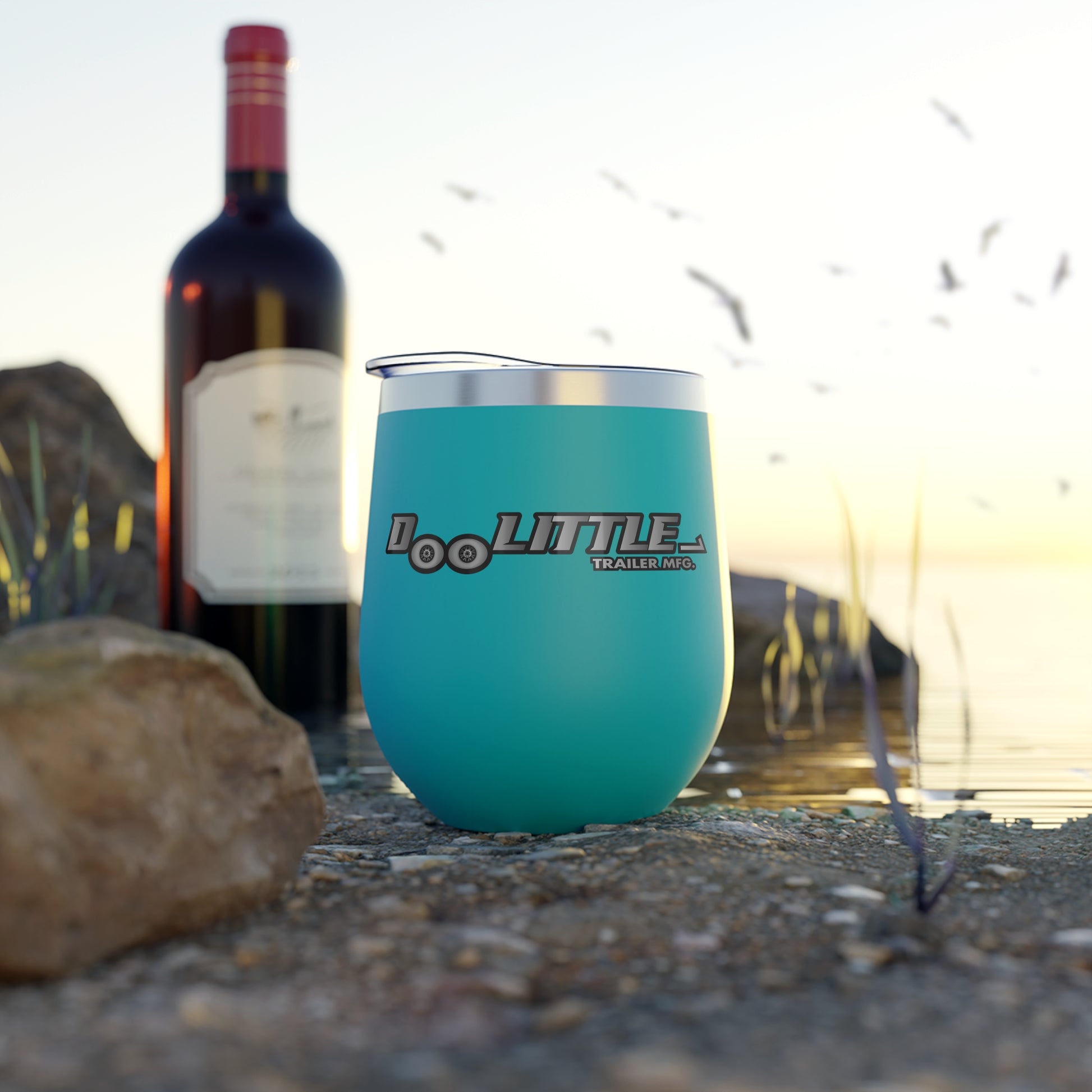 Teal wine tumbler with 'DooLittle Trailers' logo next to a bottle of red wine on a gravel surface.